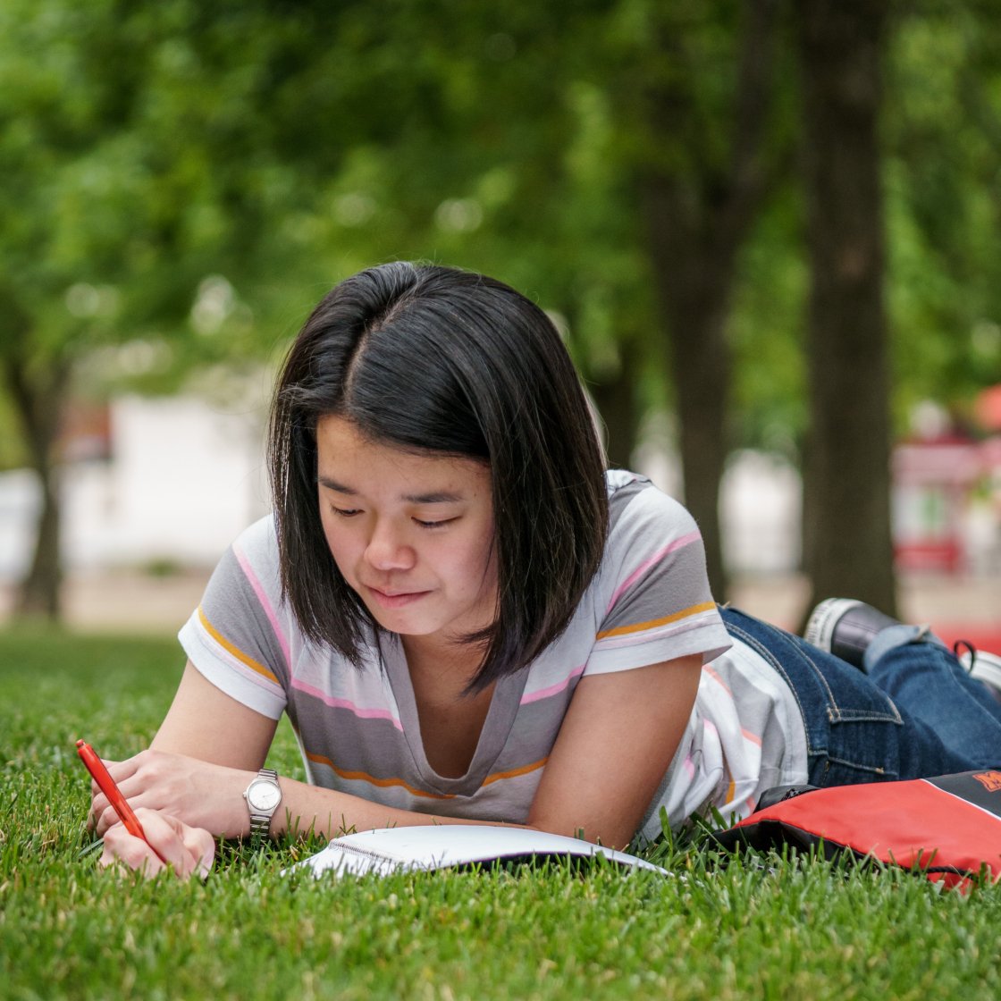 Student lying on grass, writing in a notebook