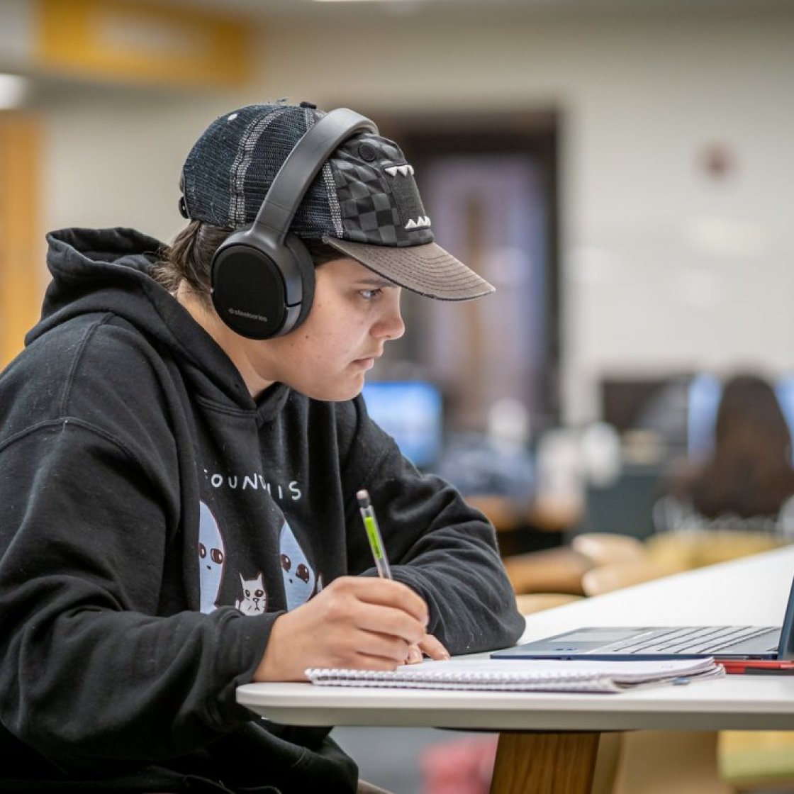 student sitting at a laptop with headphones writing in a notebook