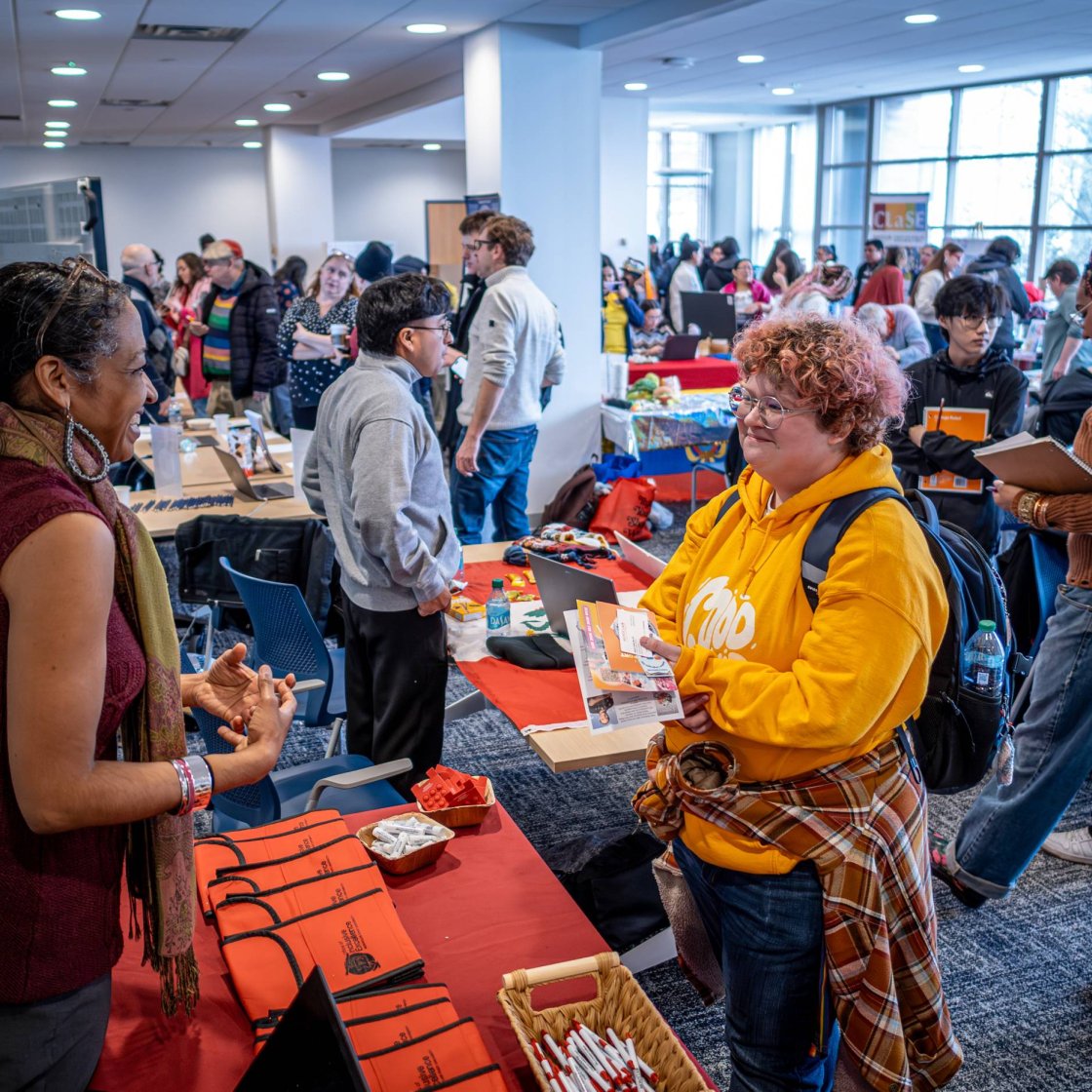Student speaking with an attendee at a table during a busy event with crowds and information tables in the background.