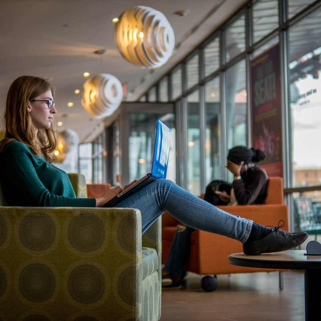 Student sitting in a lounge area, working on a laptop near large windows