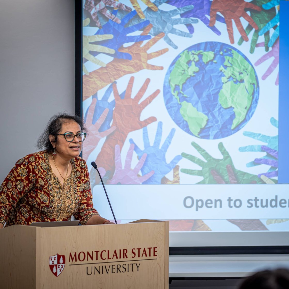 A college professor stands in front of a PowerPoint and addresses an audience