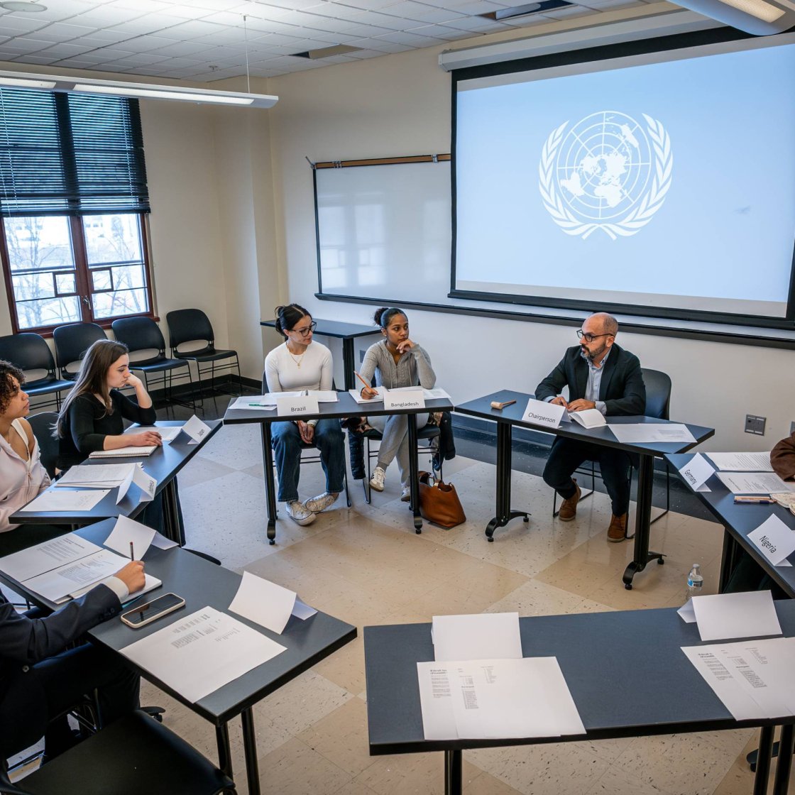 College students sit in a circle around a professor, the United Nations logo appears on the screen behind them