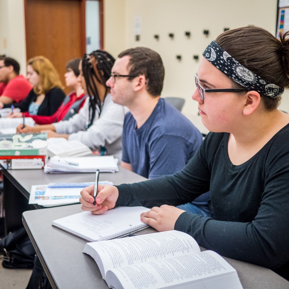 A group of students sit at desks in a classroom, writing in their notebooks
