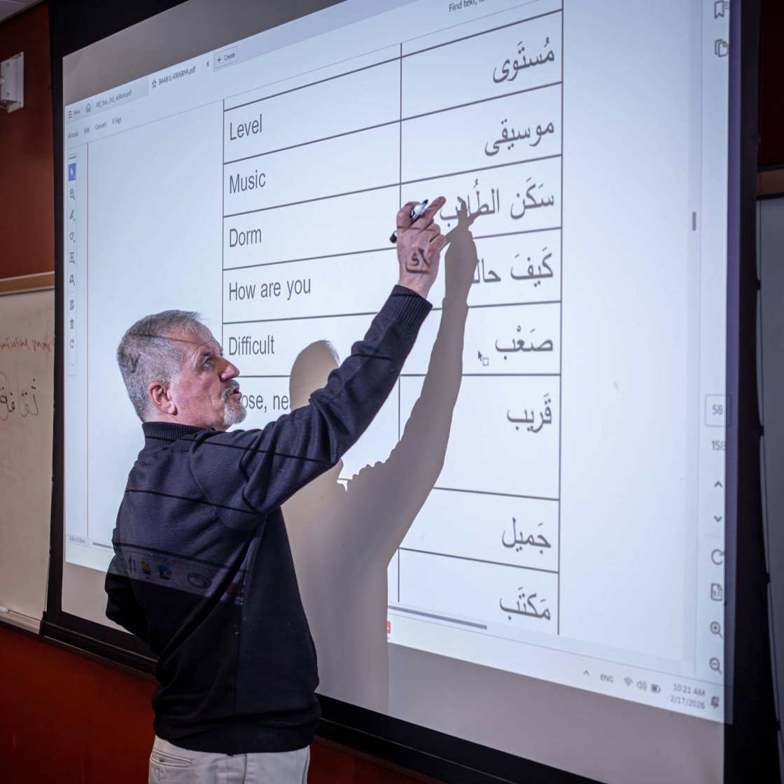 A college professor stands before a projector screen and points to something on screen 