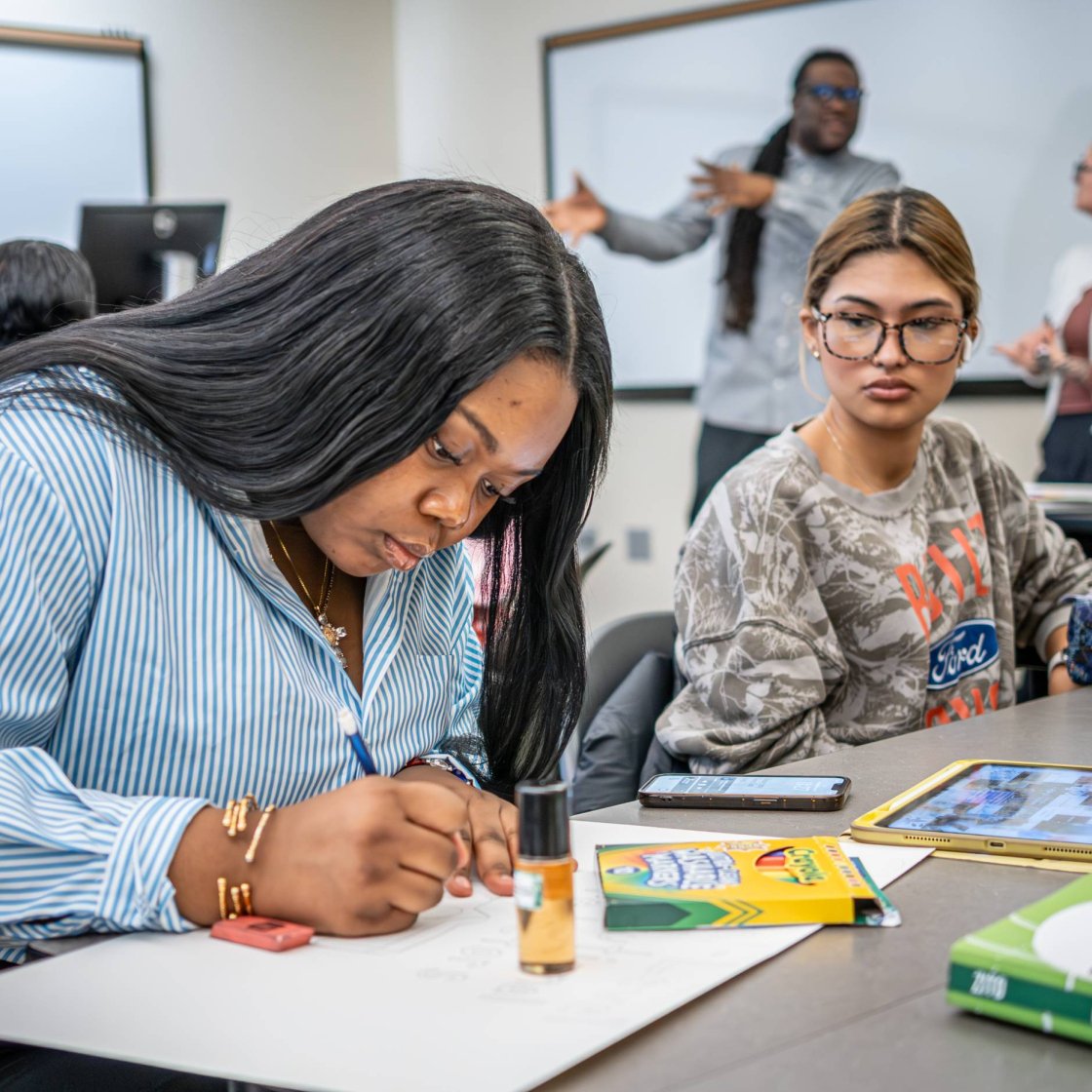 A college student works on a poster in a classroom while another student looks on