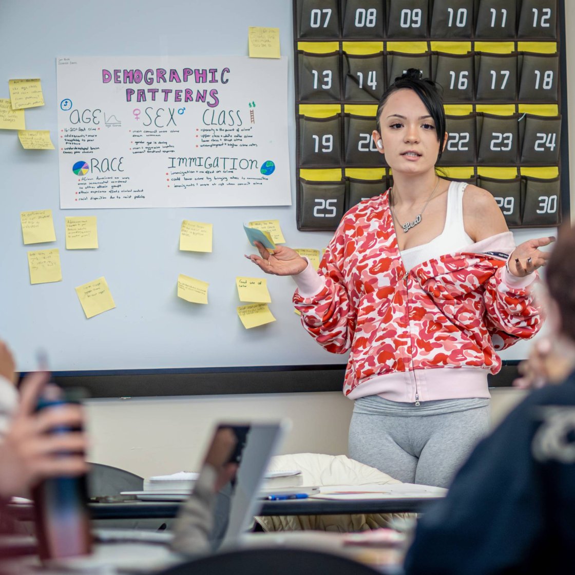 A college student points to sticky notes on a whiteboard during a presentation to a class