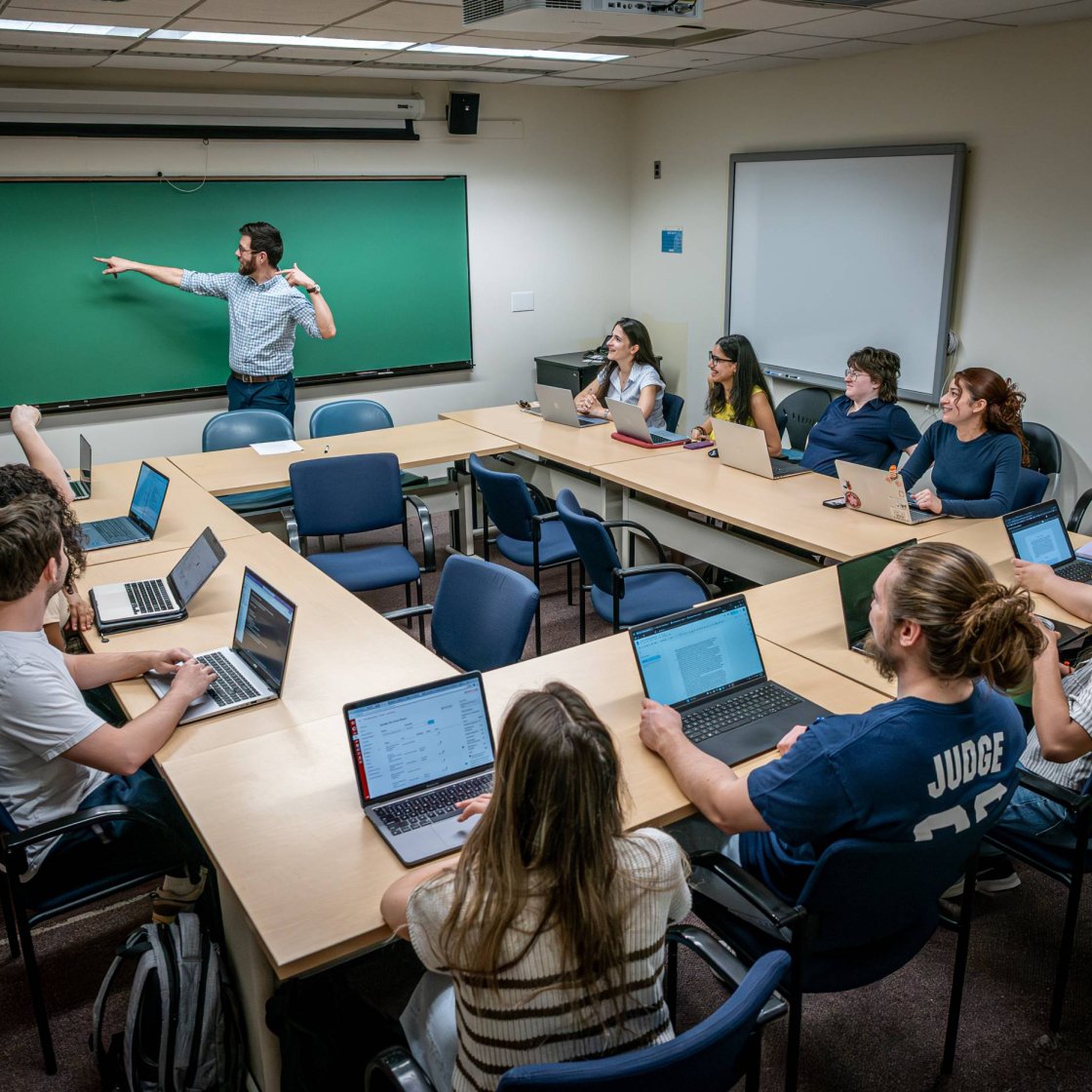 A college professor stands at a chalkboard in front of a classroom full of students