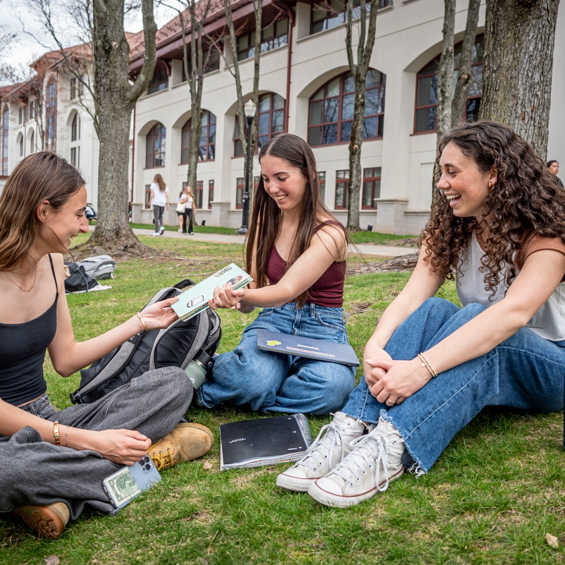 three students passing a notebook on the quad