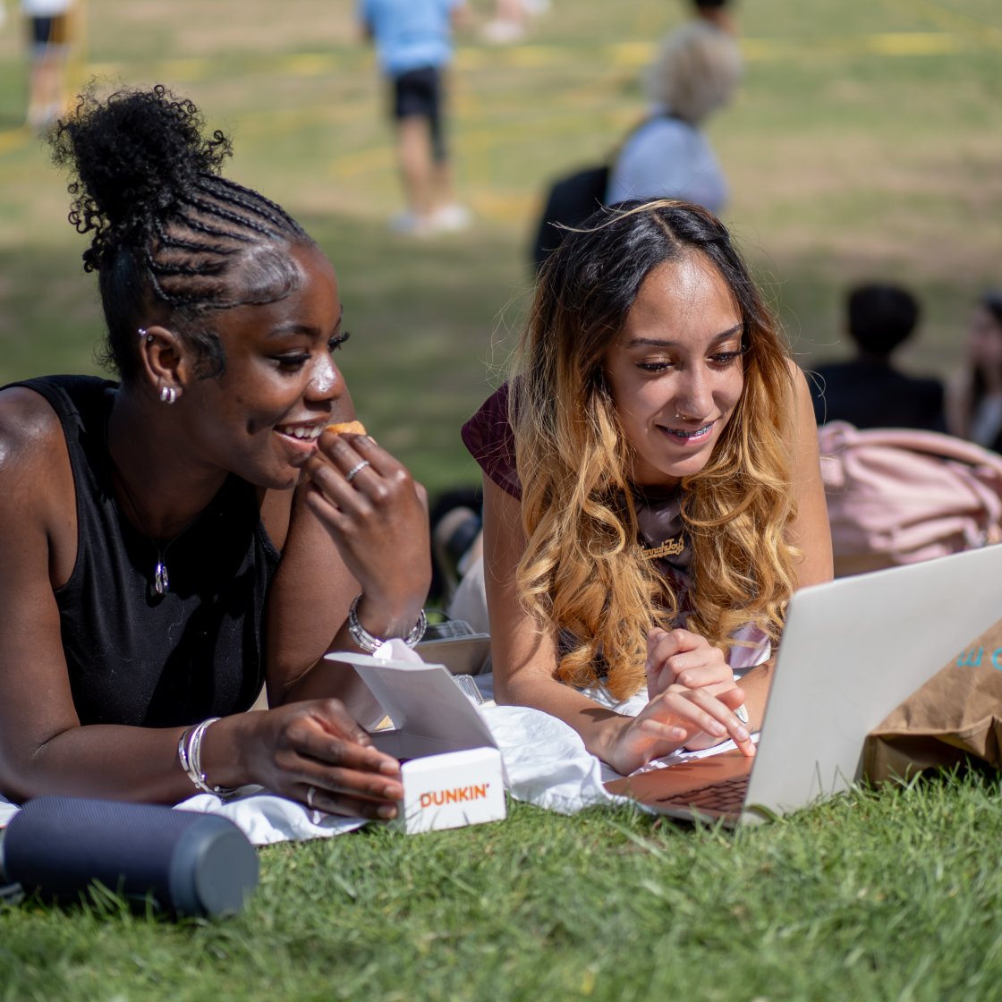 two students smiling down at laptop on quad