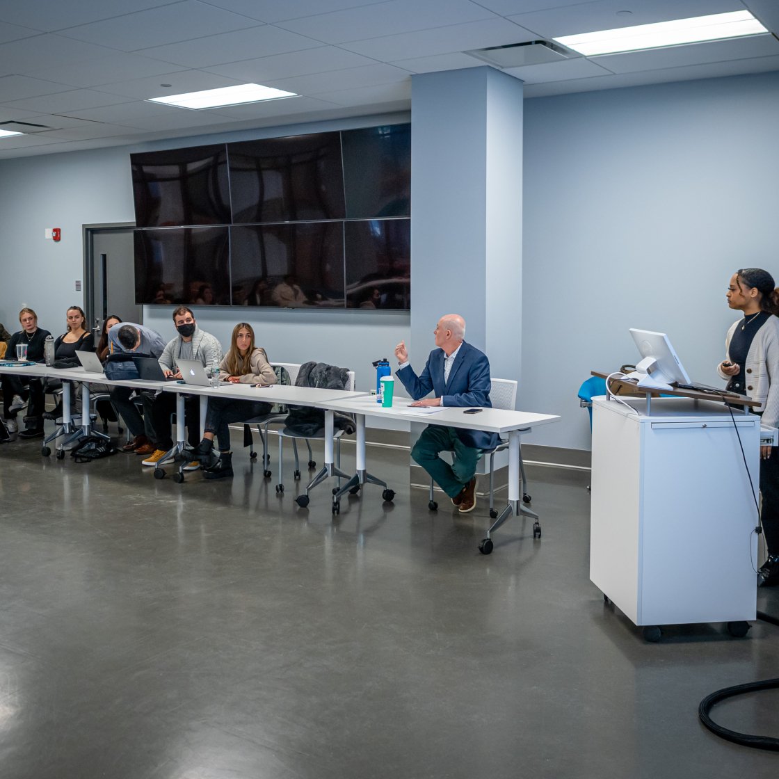 Professor speaking to the class while two stdudents on the right hand side are about to begin their presentation.