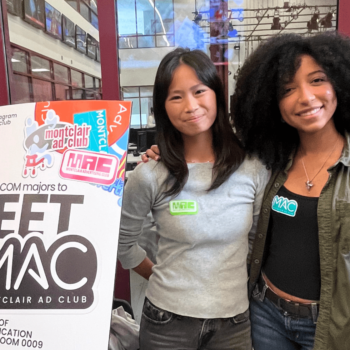Two students pose in front a Meet Montclair Ad Club recruitment poster at the club fair.