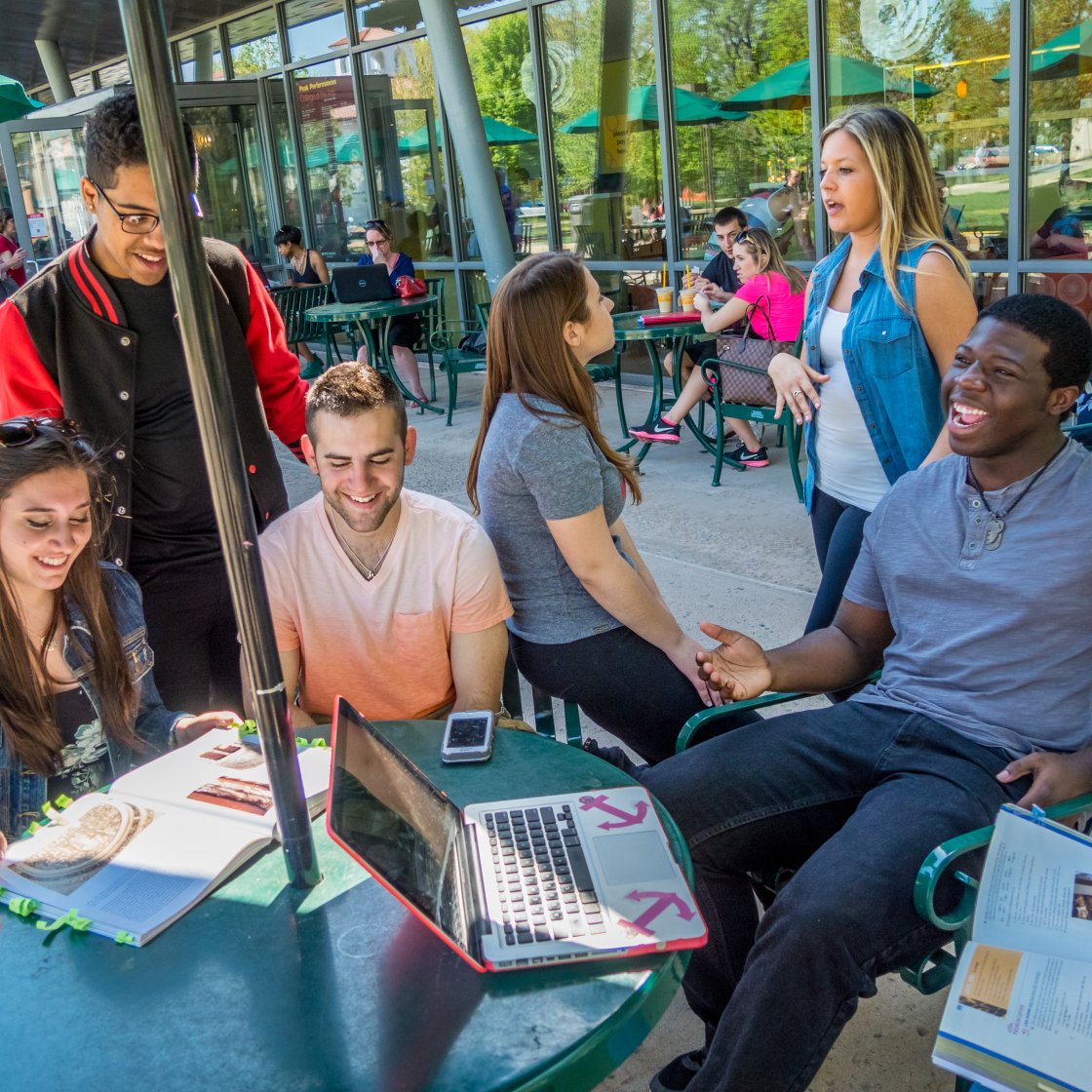 A group of college students sit around an outdoor table under an umbrella with laptops and books out