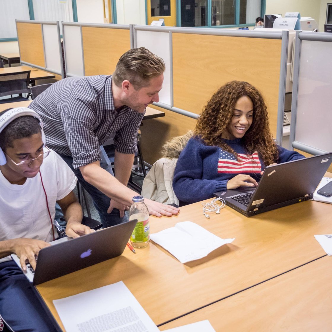 Two college students sit in front of laptops at a desk as an instructor leans over their shoulder 