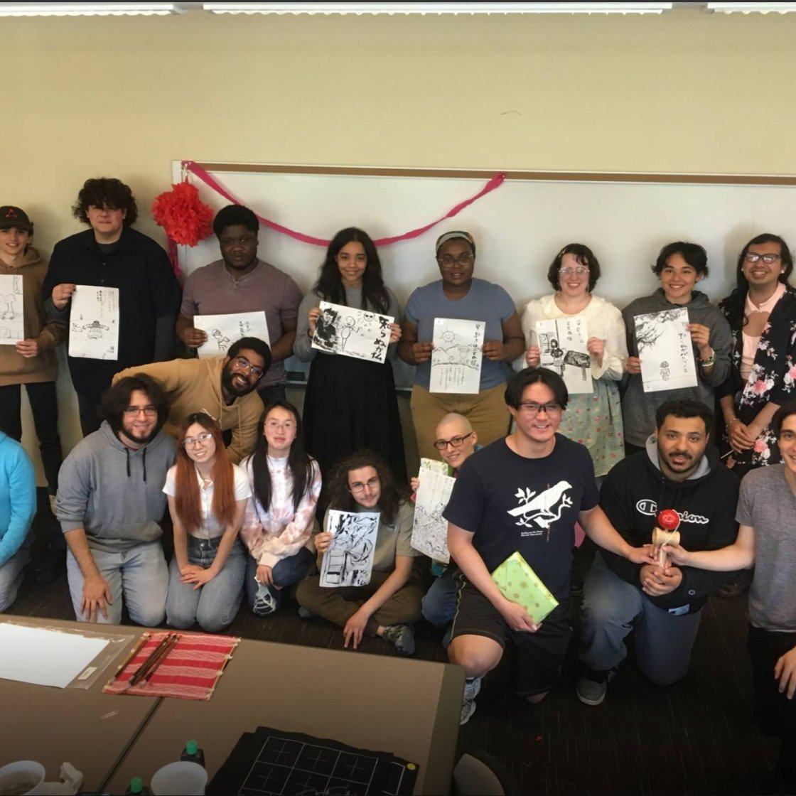 A group of college students pose in two lines in front of a whiteboard and hold up sketches they drew