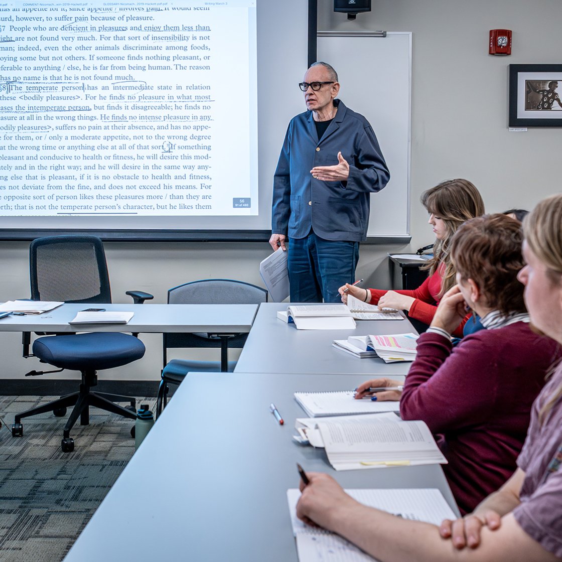 Professor Tiger Roholot in front of class speaking with screen visible next to him. Students are turned to him and listening