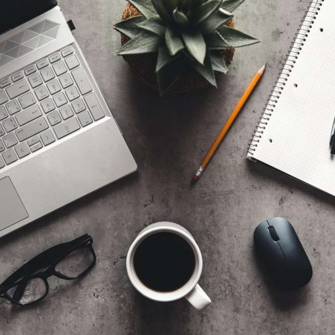 Workspace with a silver laptop, black glasses, coffee cup, wireless mouse, spiral notebook with pen, pencil, and a potted succulent.