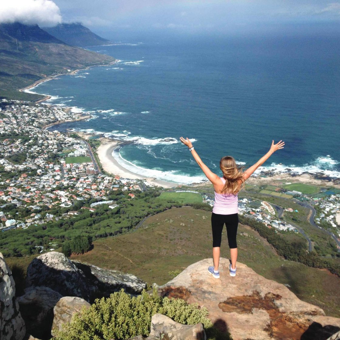 A woman stands on the edge of a tall cliff overlooking a beach