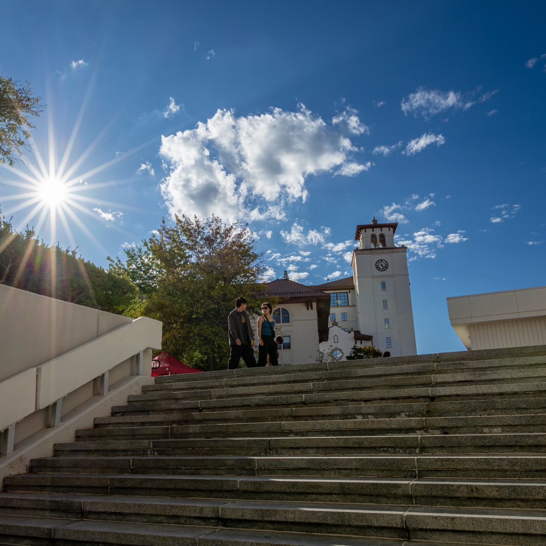 Two students walk up campus steps toward a clock tower under a bright blue sky with the sun shining through nearby trees.