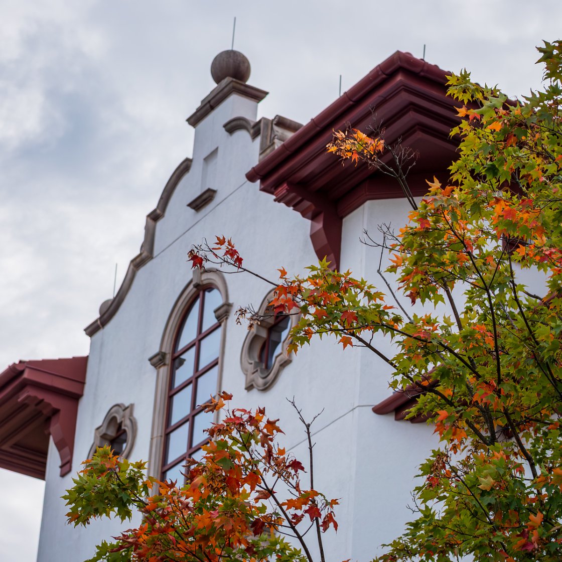 A campus building with Spanish-style architecture is framed by colorful autumn trees under a cloudy sky.