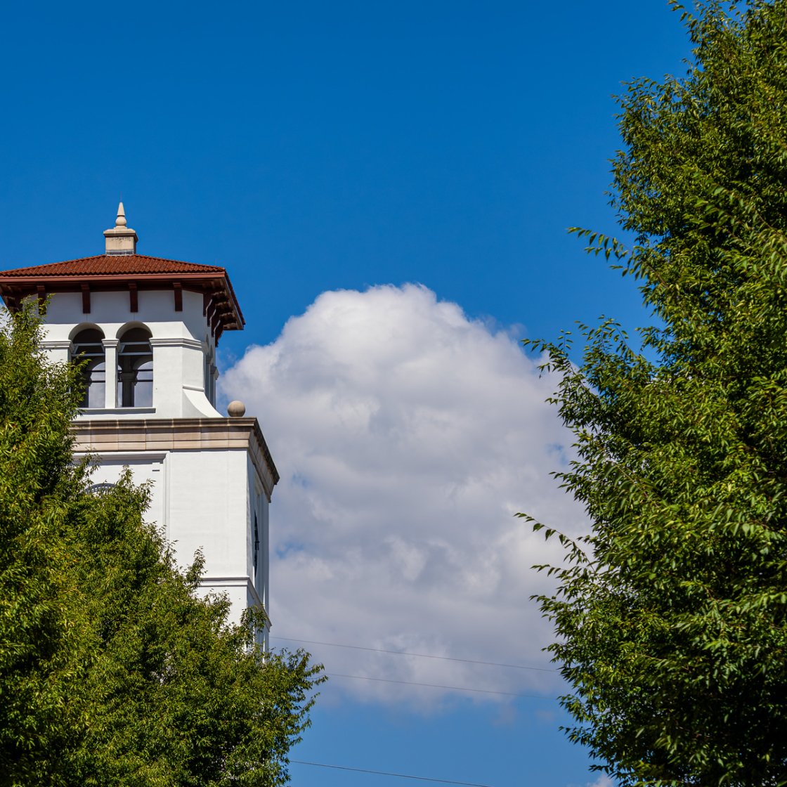 A white campus bell tower rises above green trees beneath a vivid blue sky with large white clouds.