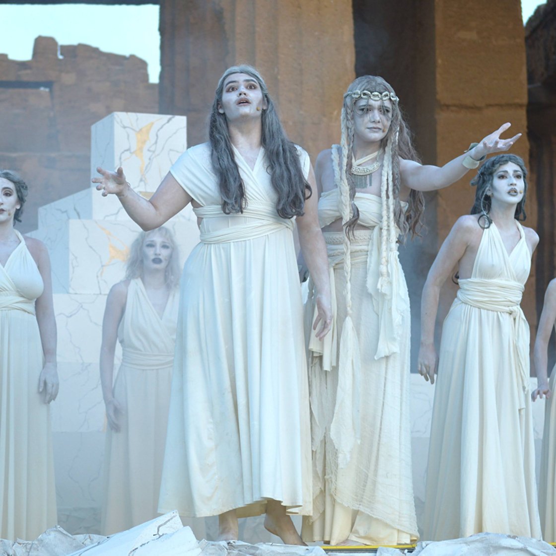 Three students dressed in white toga-style costumes performing the musical Agrigento at an ancient ruin in Italy with large stone columns in the background