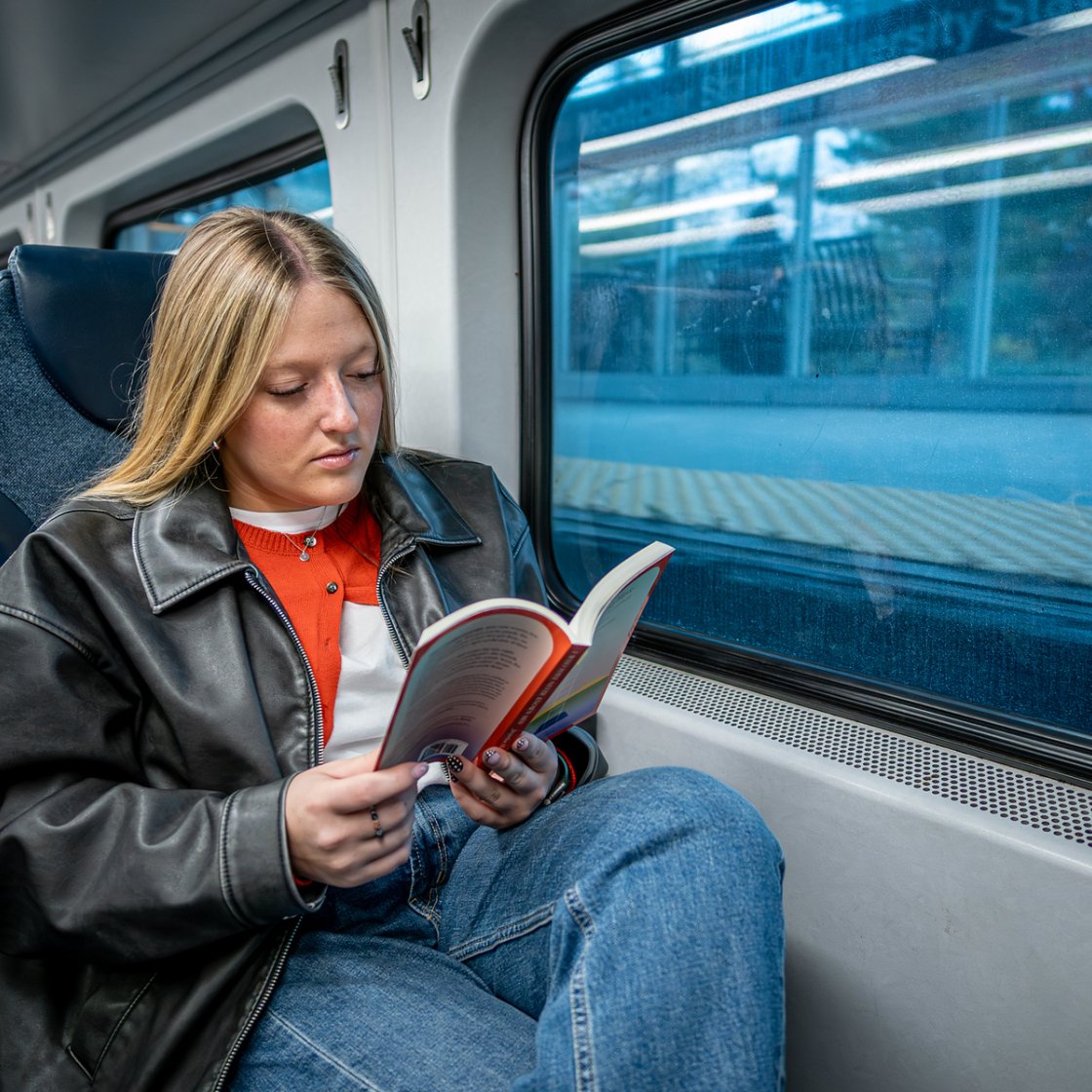 A student reads a book while seated by the window on a train, making productive use of their commute to campus.