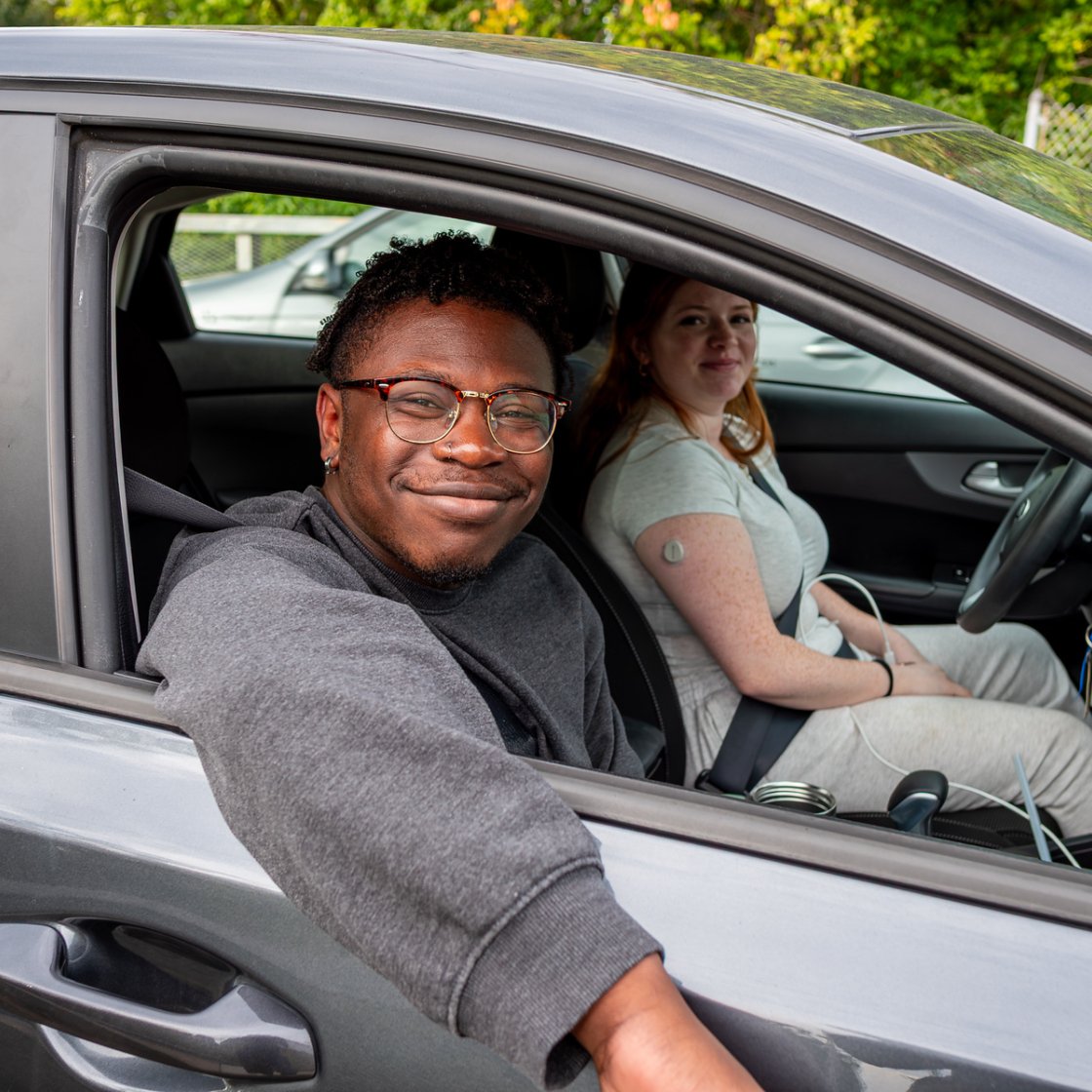 Two smiling college students sit in a parked car, with the front passenger leaning out the open window while the driver looks toward the camera.