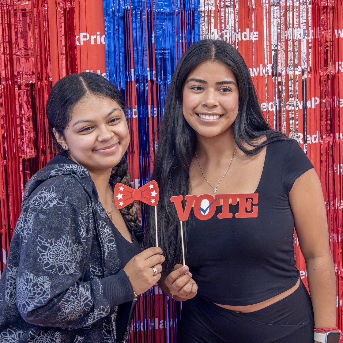 Two students pose in front of a red, white, and blue backdrop while holding voting-themed props during a campus civic engagement event.
