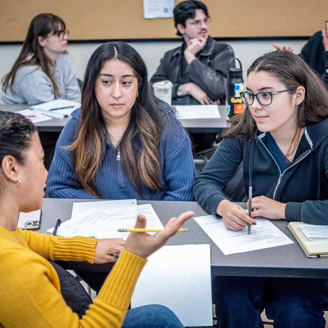 Three college students sit at a desk in a classroom and talk
