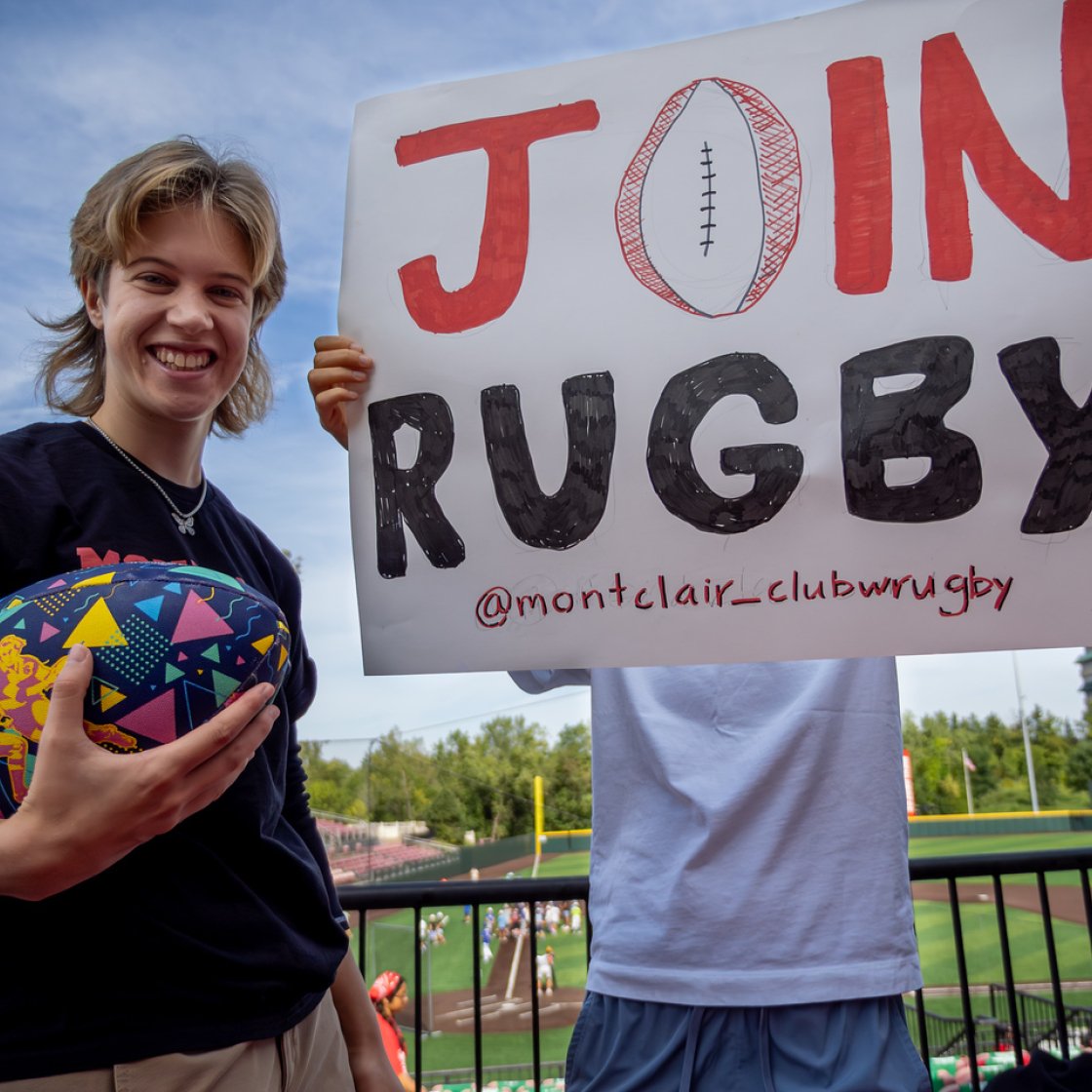 A student holding a football and the other holding up a poster "Join rugby"