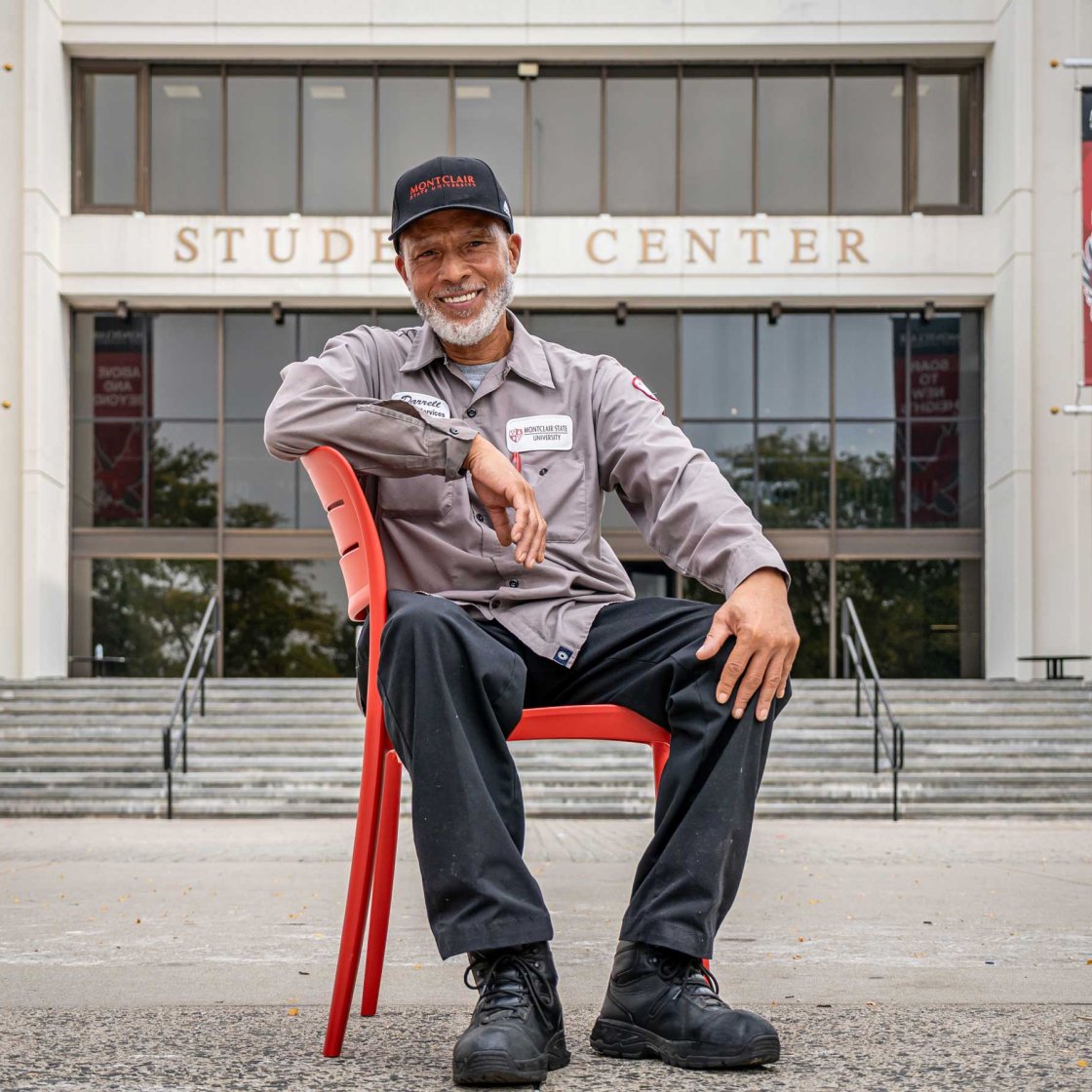 A smiling employee is seated in a red chair in front of the Student Center.