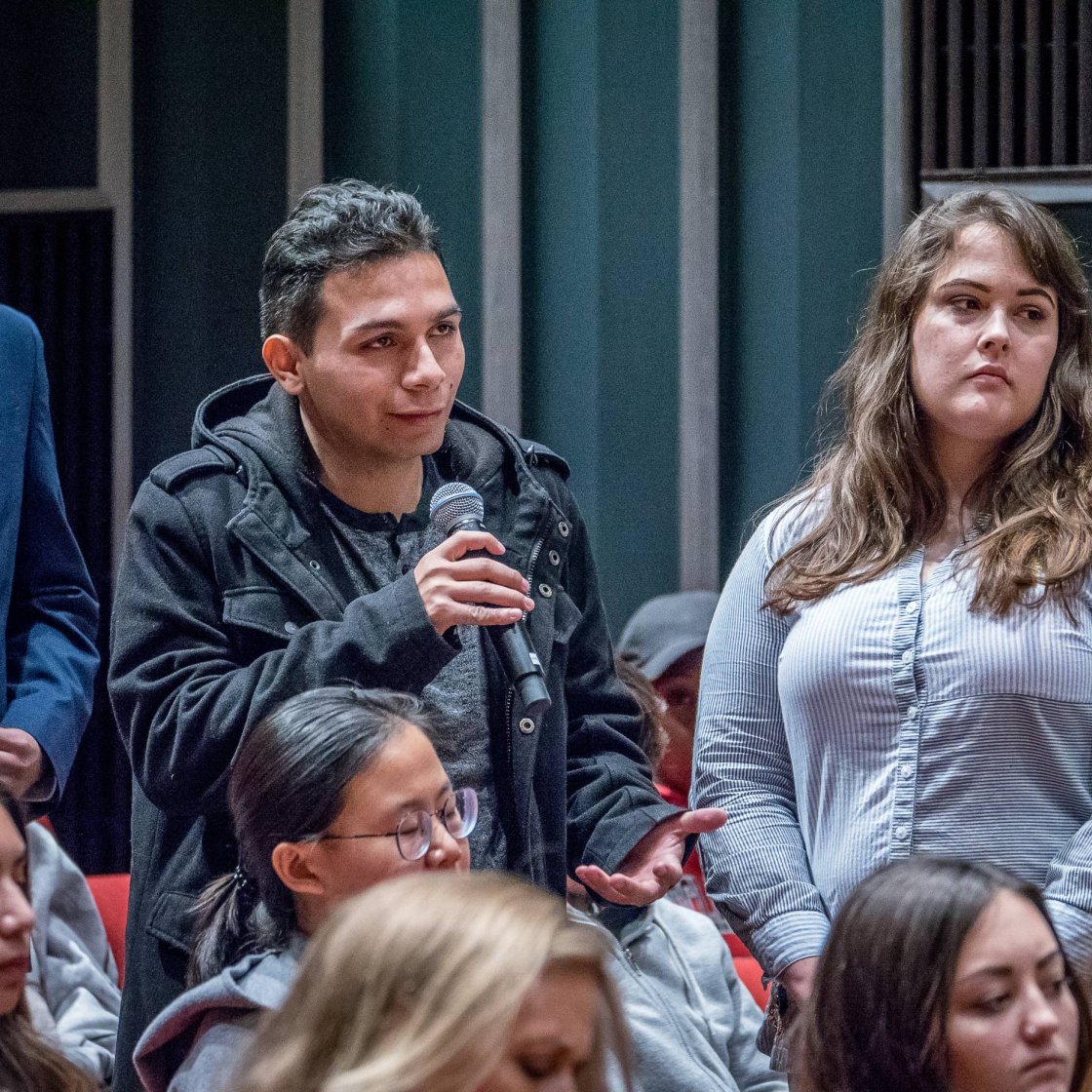 A college student stands in a crowd, holds a microphone and asks a question to a speaker