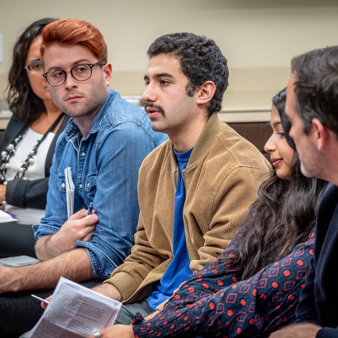 A man speaks from a seat in a classroom as two other men look on