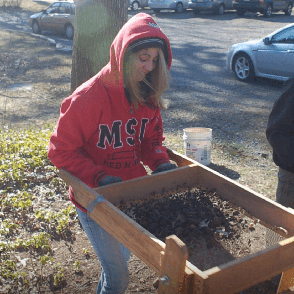 A woman in a Montclair State University University sweatshirt screens excavated soil