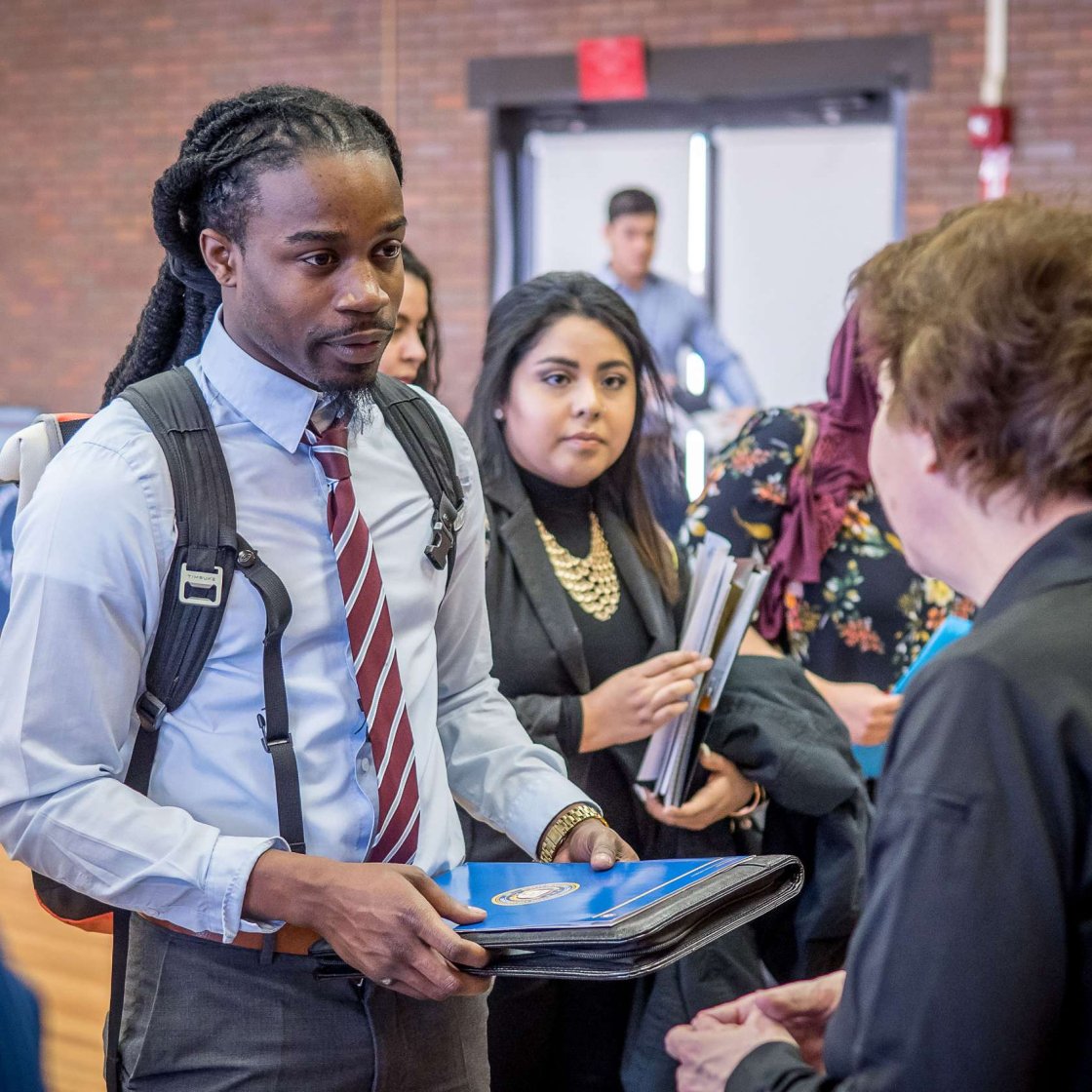 A student in shirt and tie holds portfolio while speaking with someone at a job fair