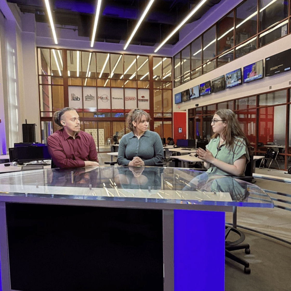 Three students sitting by the anchor desk in newsroom engaging in conversation.