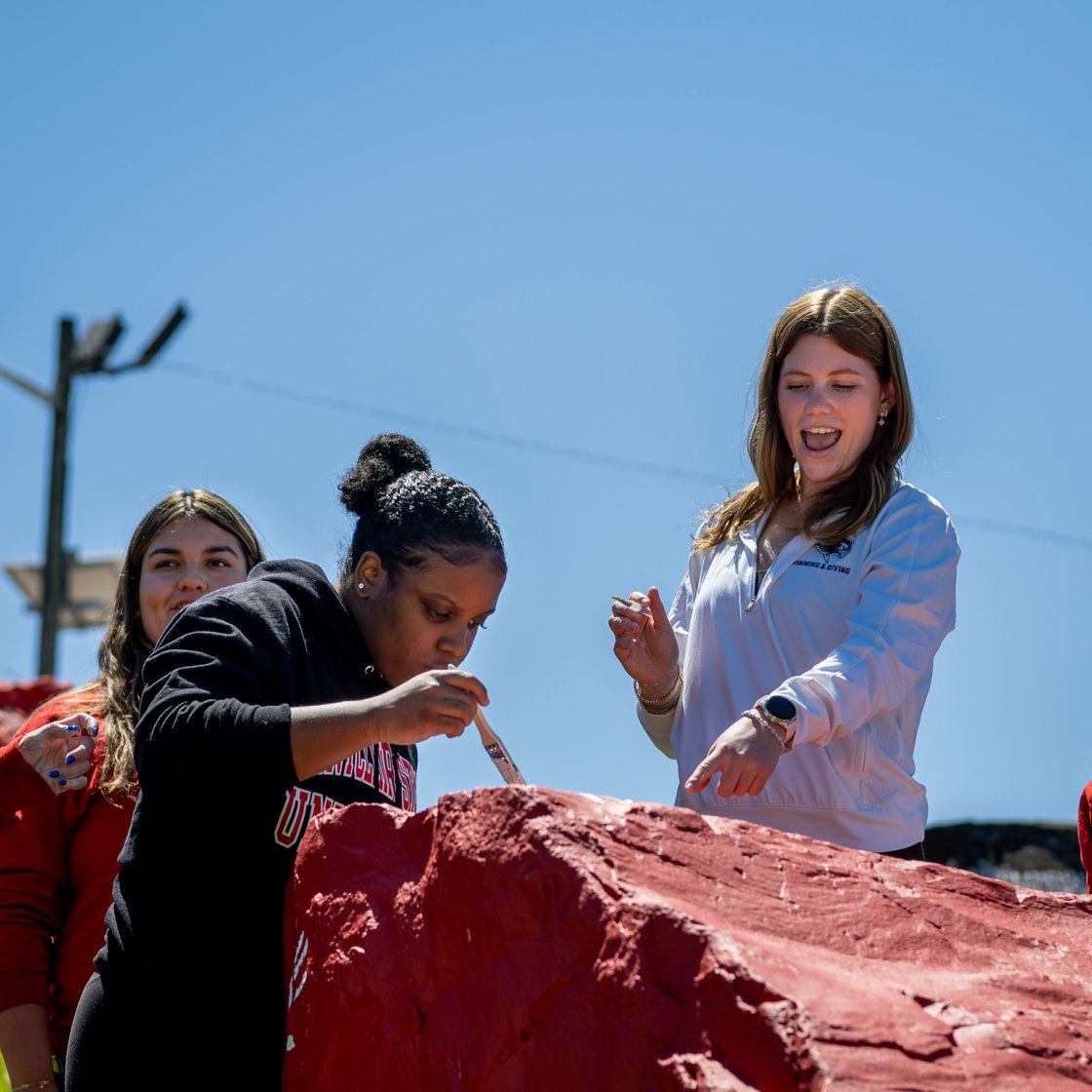 Alt text: Students work together to paint a large campus rock red during an outdoor community service activity under a clear blue sky.