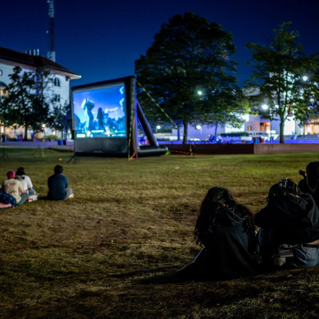 Students sit on the campus lawn at night watching an outdoor movie projected on a large inflatable screen.