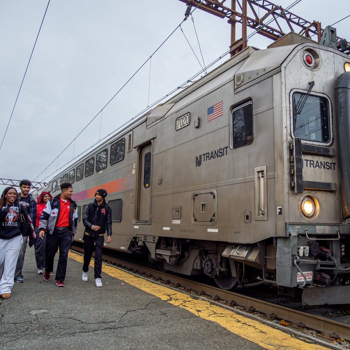Students walk along the platform beside a stopped NJ TRANSIT train at a station on an overcast day.
