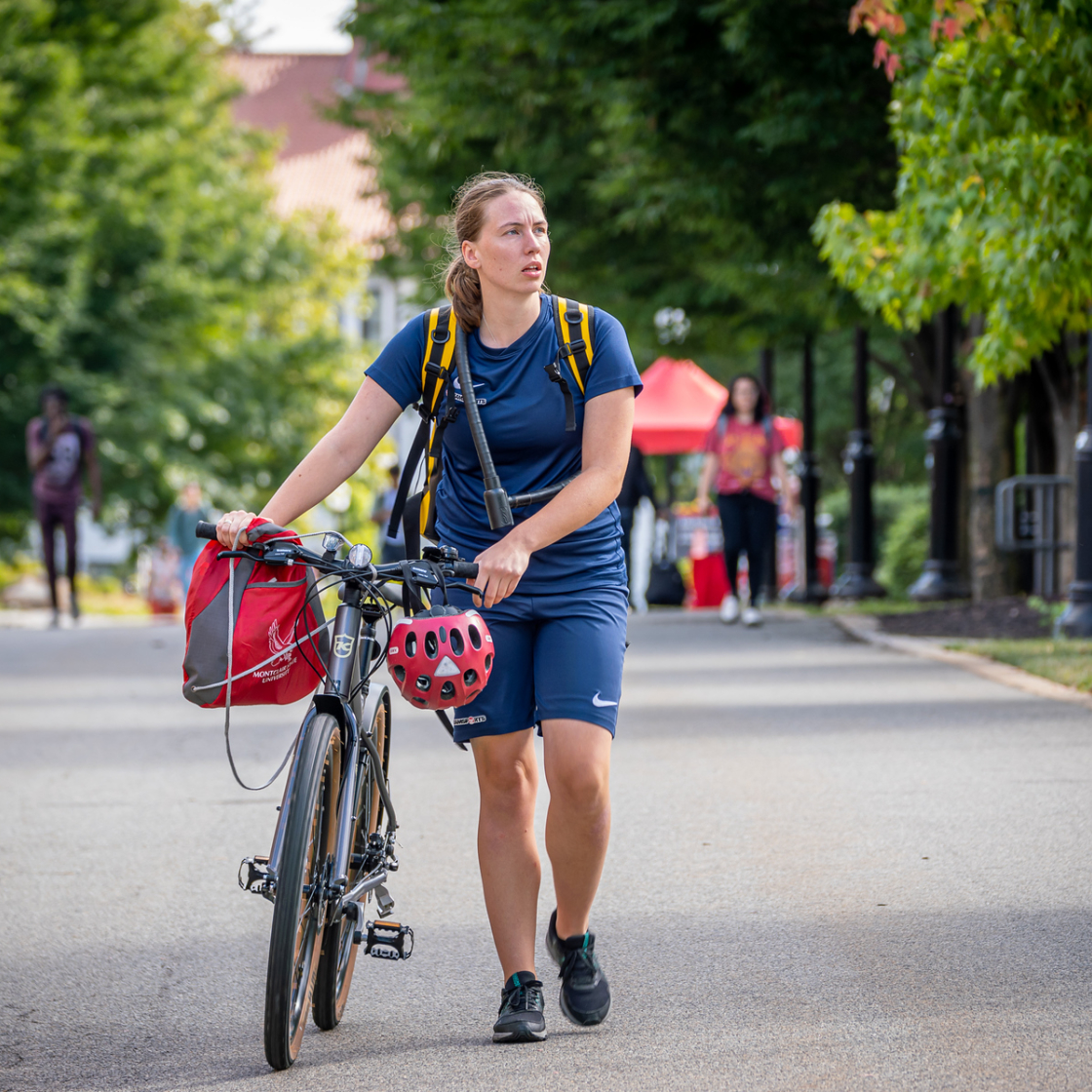 A student walks a bicycle with a red Montclair University bag and helmet attached while traveling along a tree-lined campus pathway.