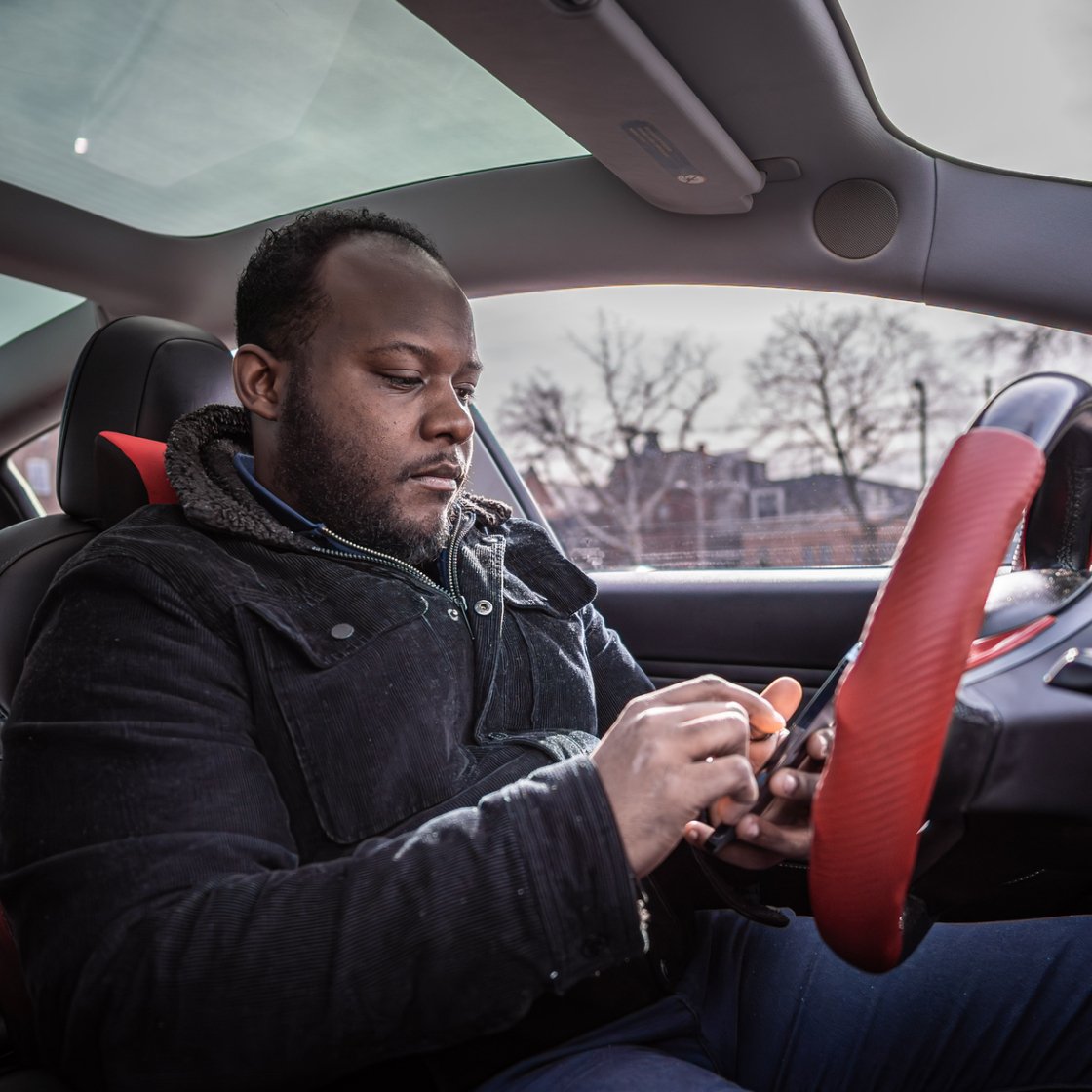 A student sits in the driver's seat of a parked car using a smartphone before starting their trip.