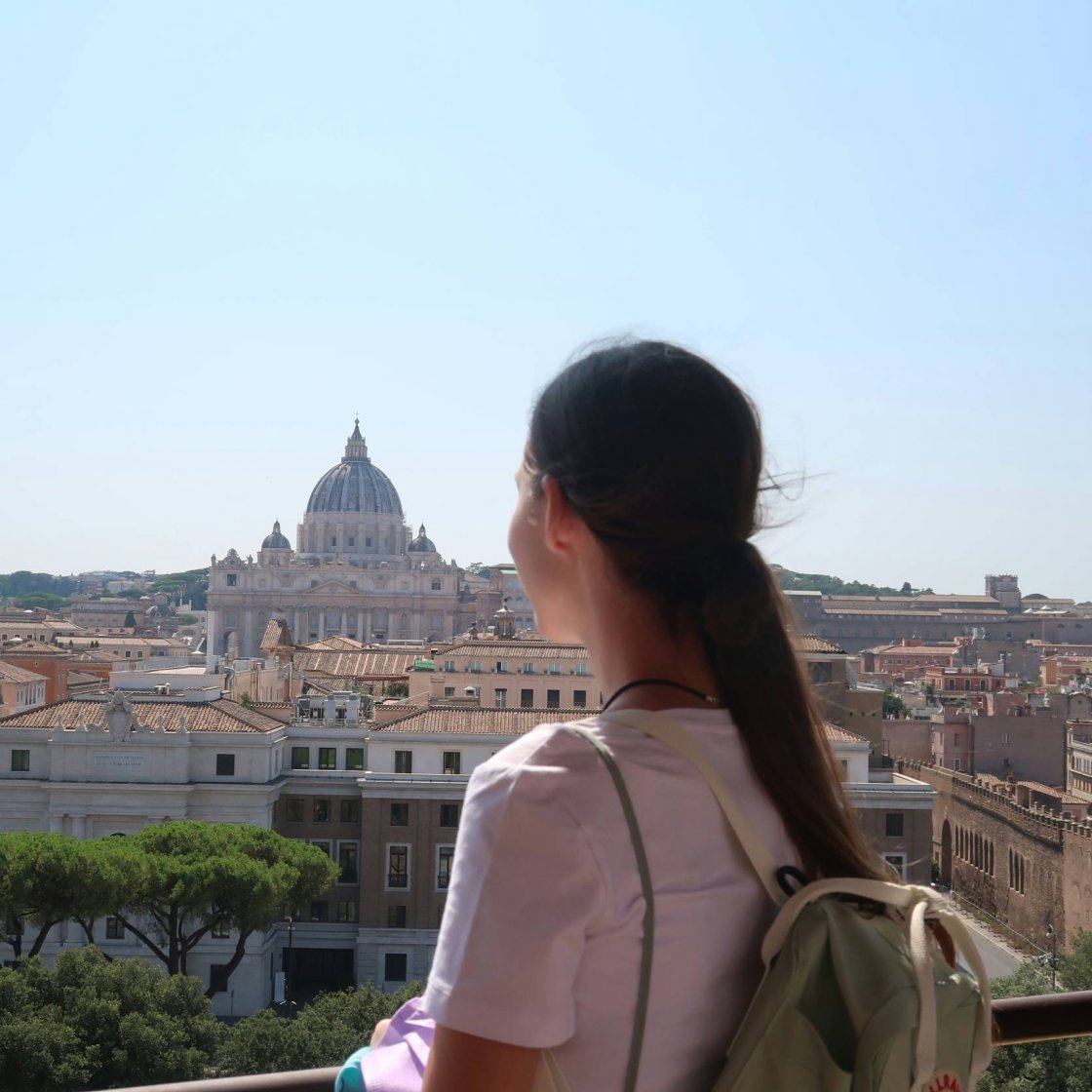 A young woman wearing a backpack stands at a railing over looks out over Rome