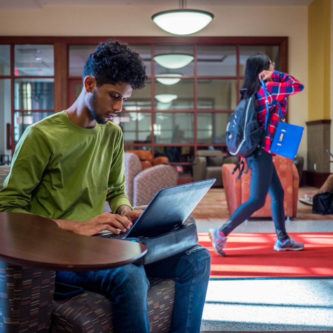 A student sits at a table working on his laptop next to a busy hallway. 