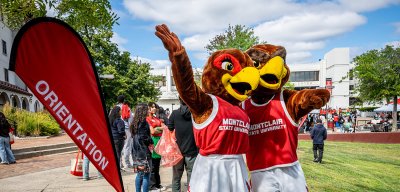 Mascots Roxy and Rocky standing outside with by the Orientation flag