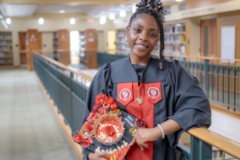 Lataiva Balmer, a psychology major and member of the first graduating class of Bloomfield College of Montclair State University, is shown in the college library wearing her cap and gown on the Bloomfield campus. She holds a decorated graduation cap.