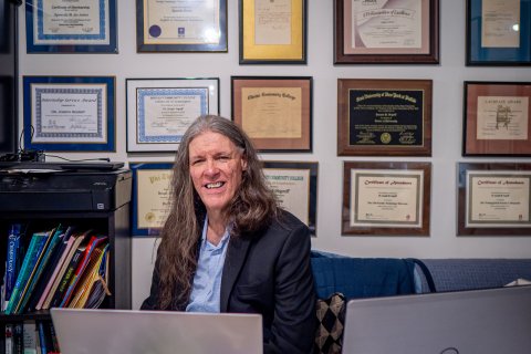 A man sits at his desk smiling with many diplomas on the wall behind him.