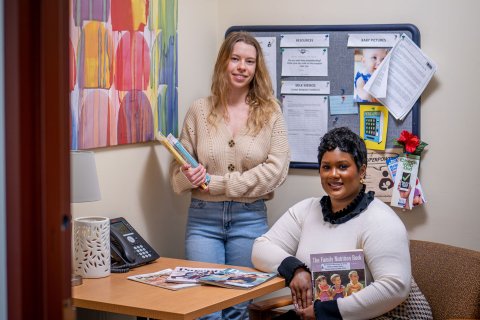 One woman stands and another sits while holding books.