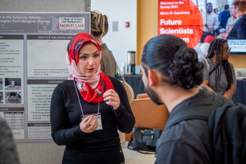 Busra Anil, a Montclair State University student wearing a red and white patterned hijab and black sweater, speaks with a guest during the Spring 2024 Mario M. Casabona Future Scientists Competition. She stands in front of her research poster on carbon sequestration, explaining her findings.