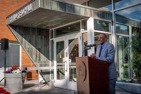 A man in a suit stands at a podium in front of the Irvin D. Reid Hall entrance.