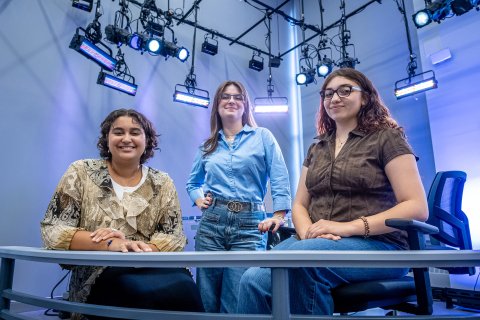In a newsroom, three women, two seated at a desk and one standing between them pose with studio lights shining behind them.
